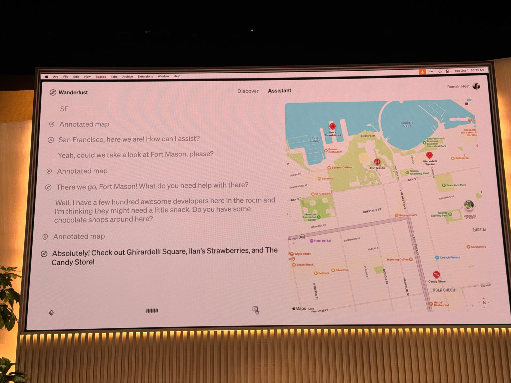 The image shows a large display screen mounted on a wall, showcasing a map application called Wanderlust. The screen has a pink background with a map of San Francisco on the right side.
The top of the screen shows macOS menu bar with icons and the time Tue Oct 1 10:35 AM.
The left side of the screen displays a conversation:
SF
Annotated map
San Francisco, here we are! How can I assist?
Yeah, could we take a look at Fort Mason, please?
Annotated map
There we go, Fort Mason! What do you need help with there?
Well, I have a few hundred awesome developers here in the room and I'm thinking they might need a little snack. Do you have some chocolate shops around here?
Annotated map
Absolutely! Check out Ghirardelli Square, Ilan's Strawberries, and The Candy Store!
The right side shows a map of San Francisco, focusing on the Fort Mason area. Various landmarks and streets are labeled, including Aquatic Park, Ghirardelli Square, and Fisherman's Wharf.
At the bottom of the screen, there are icons for microphone input, text input, and map layers.
The screen is mounted above what appears to be wood paneling, and a small portion of a plant is visible in the lower-left corner of the image.