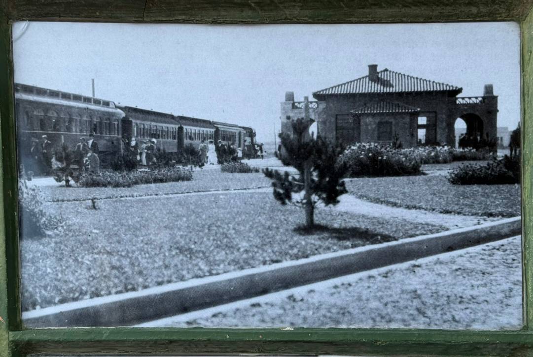 Historical black and white photograph showing a train station with a steam train on the left and a Spanish-style station building with arched entrances on the right. It's clearly the same building, though the modern one has had a bunch of extra extensions added to it and doesn't look nearly as much like a train station.