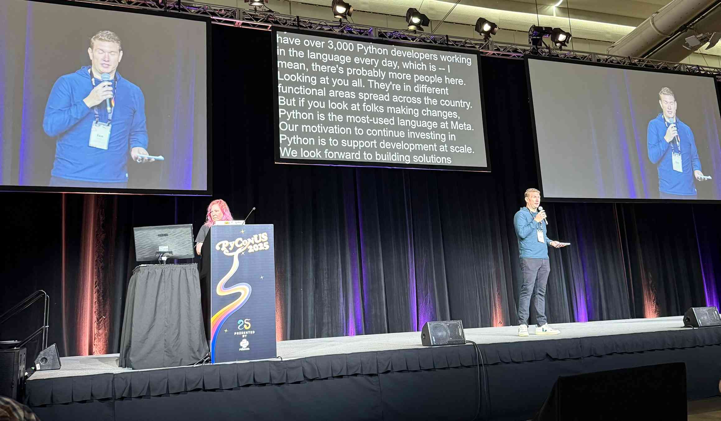 Conference presentation at PyCon US 2025 showing speaker on stage in blue shirt with large screens displaying his image and slide text: "have over 3,000 Python developers working in the language every day, which is -- I mean, there's probably more people here. Looking at you all. They're in different functional areas spread across the country. But if you look at folks making changes, Python is the most-used language at Meta. Our motivation to continue investing in Python is to support development at scale. We look forward to building solutions"