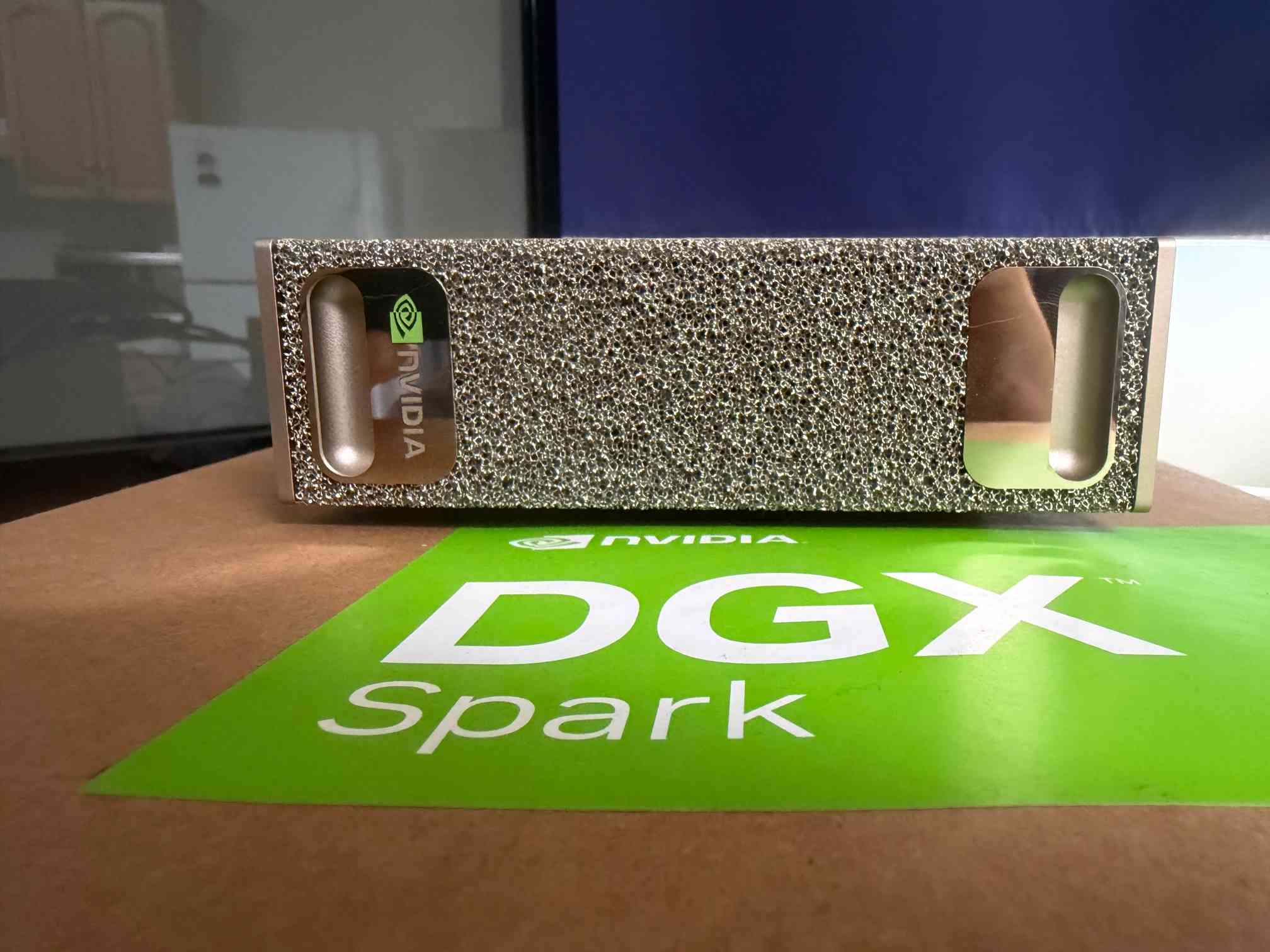 A rectangular small computer, sitting horizontally on a box. It is about the width of a Mac Mini. It has a NVIDIA logo on  a reflective handle portion, then textured silver metal front, then another reflective handle at the other end. It's pretty and a bit weird looking. It sits on the box it came in, which has NVIDIA DGX Spark written on it in white text on green.