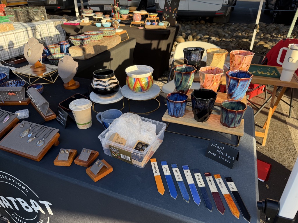 Outdoor craft market booth displaying handmade ceramics and jewelry on a navy tablecloth with "NATBAT CREATIONS CALIFORNIA USA" logo. Items include colorful glazed ceramic cups in blue, orange, and black; decorative bowls including a rainbow-striped piece; jewelry pendants and earrings on wooden display stands; ceramic plant markers in various colors labeled "Artichoke", "Cilantro", "Chili", "Oregano", "Potato", "Pumpkin", "Sage".