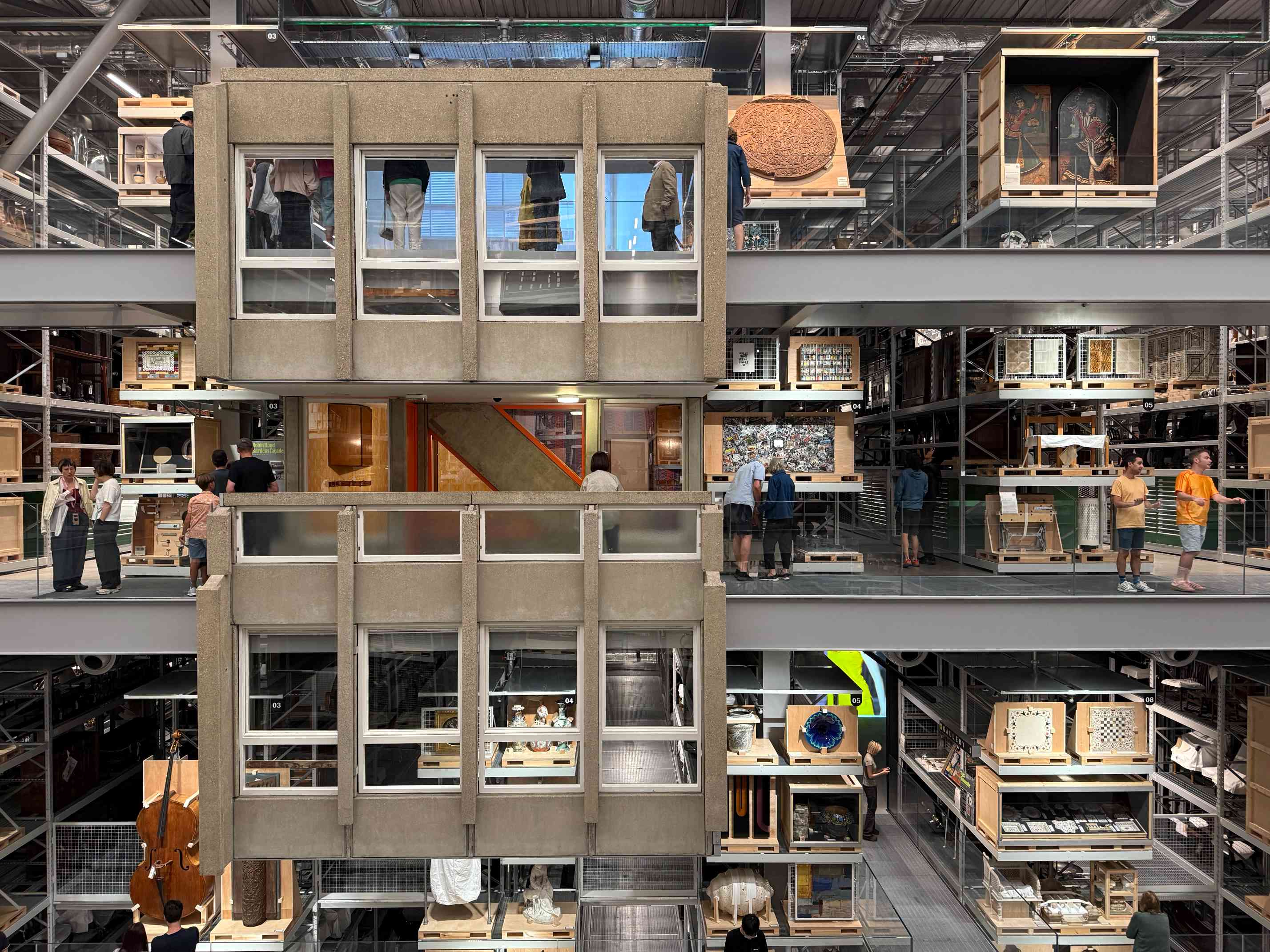 Three levels of the Storehouse, each with walkways full of people looking at a variety of exhibits on shelves. Two huge concrete facades from the Robin Hood Gardens hang between the floors.