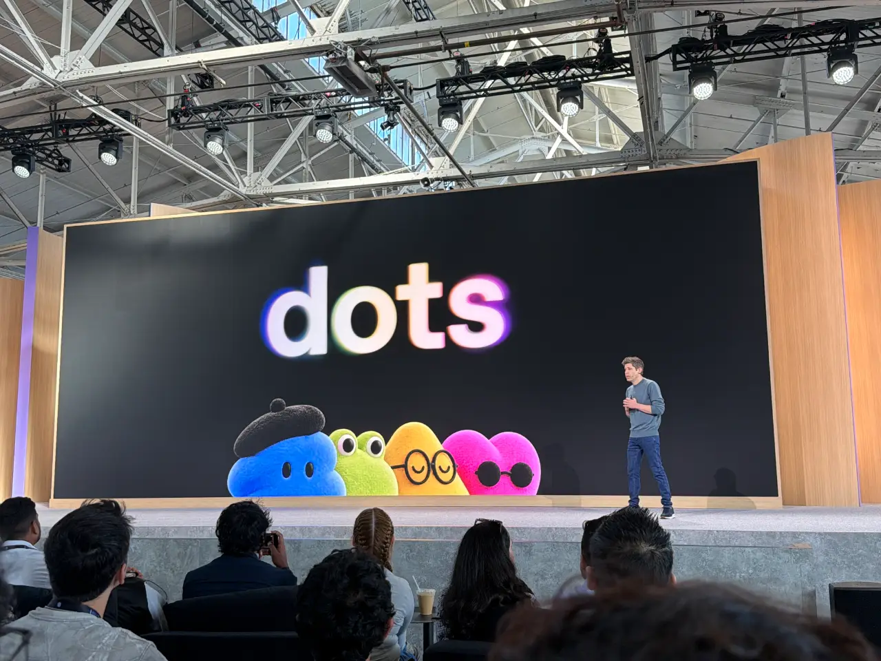 A presenter stands onstage before an audience beside a giant screen displaying “dots” above four colorful cartoon characters. The event is in a large indoor venue with exposed roof trusses and stage lights.