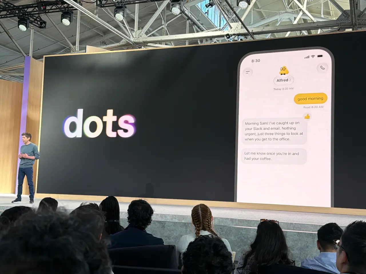 A presenter stands before an audience onstage beside a large screen displaying the “dots” logo and a mock phone conversation with an assistant named Alfred. The messages read: “good morning,” “Morning Sam! I’ve caught up on your Slack and email. Nothing urgent, just three things to look at when you get to the office,” and “Let me know once you’re in and had your coffee.”