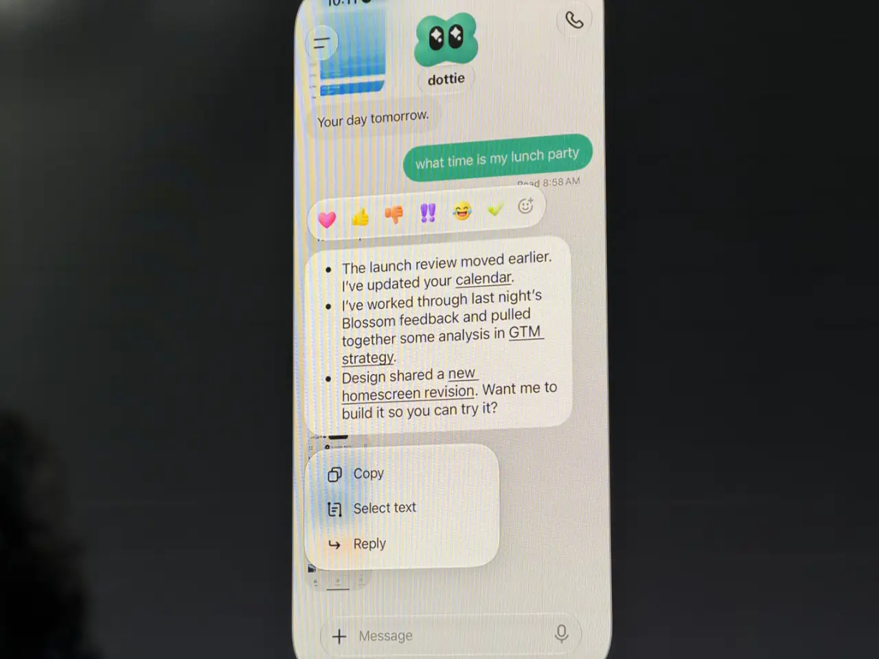 A close-up of a smartphone shows a chat with an assistant named “dottie.” After the user asks, “what time is my lunch party,” the assistant lists work updates: a launch review moved earlier, calendar updated, analysis of Blossom feedback for GTM strategy, and a new homescreen revision; a menu with “Copy,” “Select text” and “Reply” is open.