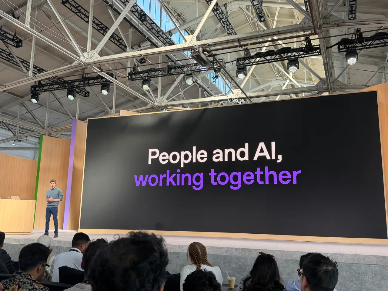 Sam Altman stands on a conference stage before a seated audience, beside a large screen reading “People and AI, working together.”