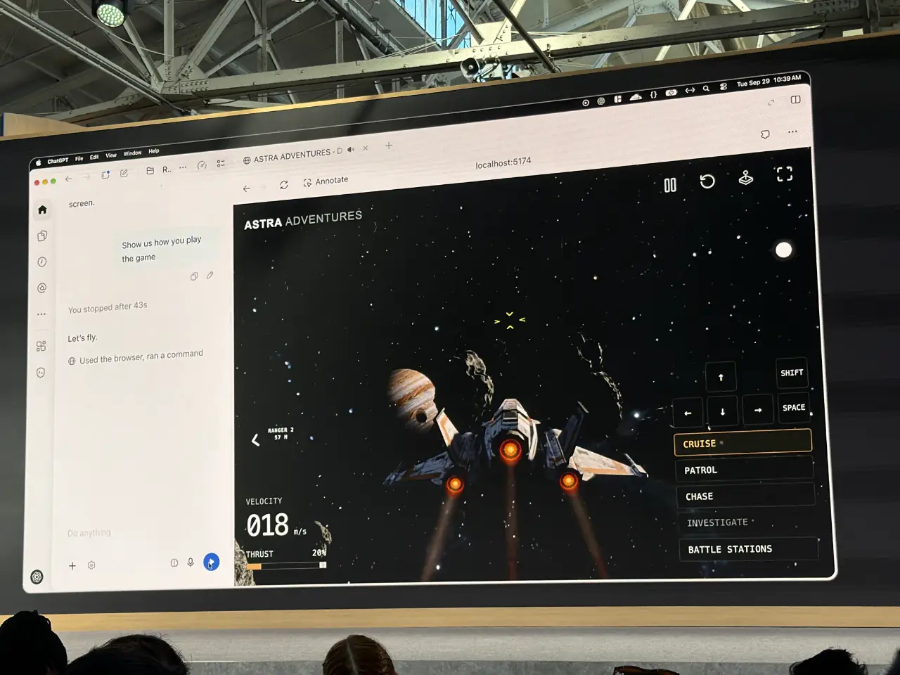 A large screen in an industrial event space shows a space-flight game called “ASTRA ADVENTURES,” with a spacecraft approaching asteroids and a planet. The interface shows velocity 018 m/s, thrust 20%, and controls including “CRUISE,” “PATROL,” “CHASE,” “INVESTIGATE” and “BATTLE STATIONS”; audience members’ heads are visible along the bottom.