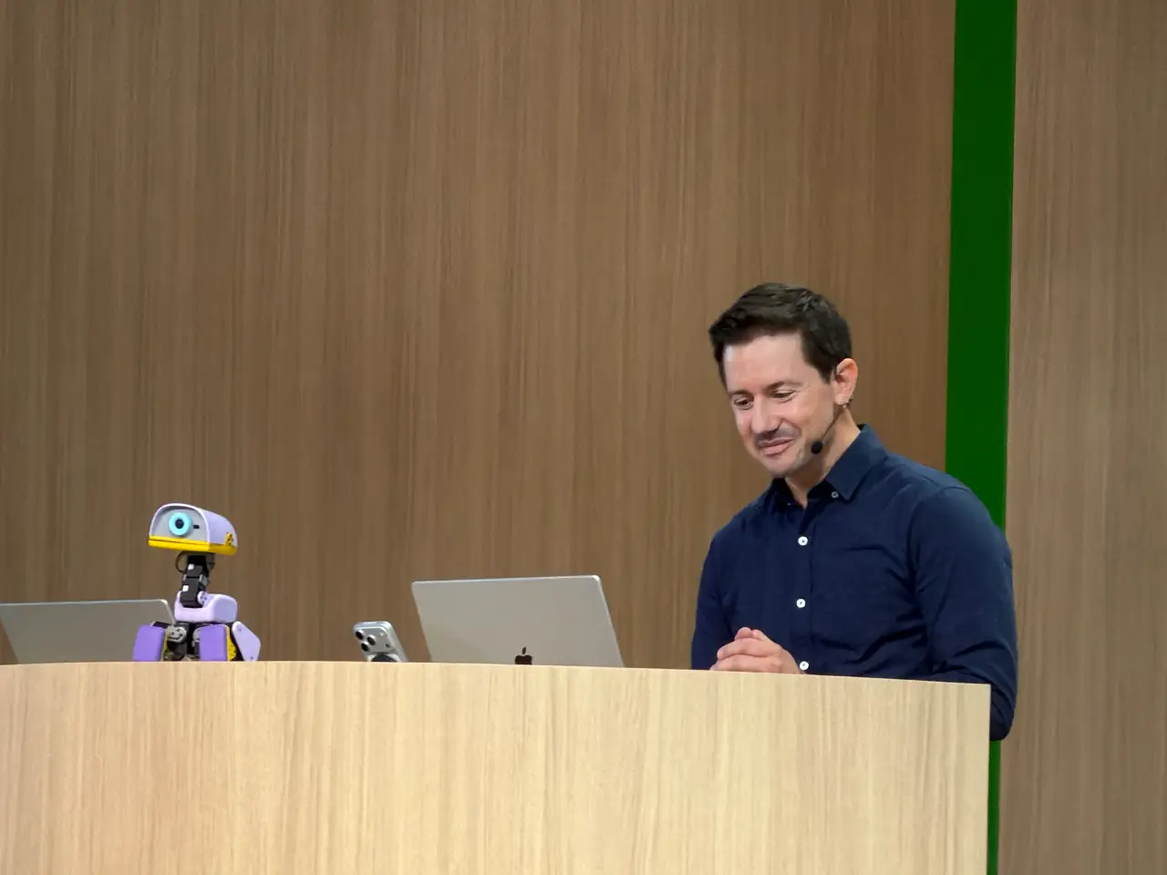 A man wearing a headset microphone stands behind a wooden lectern beside a small purple-and-yellow robot, with laptops on the lectern. A wood-paneled wall with a vertical green stripe forms the backdrop.