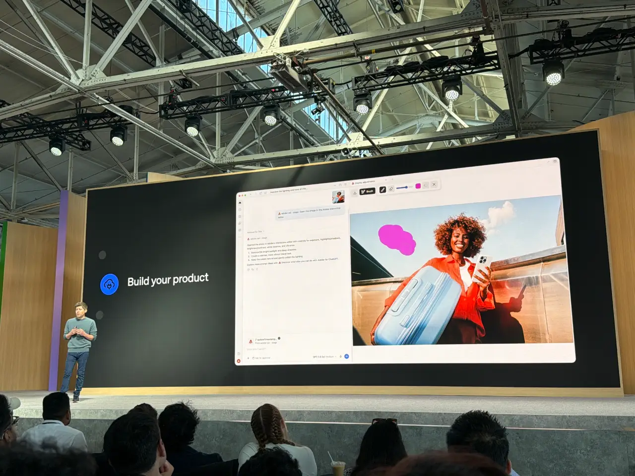 A presenter stands onstage at a technology event beside a large screen headed “Build your product.” The screen shows a software interface generating or editing an image of a smiling woman with a suitcase, while audience members watch from the foreground.