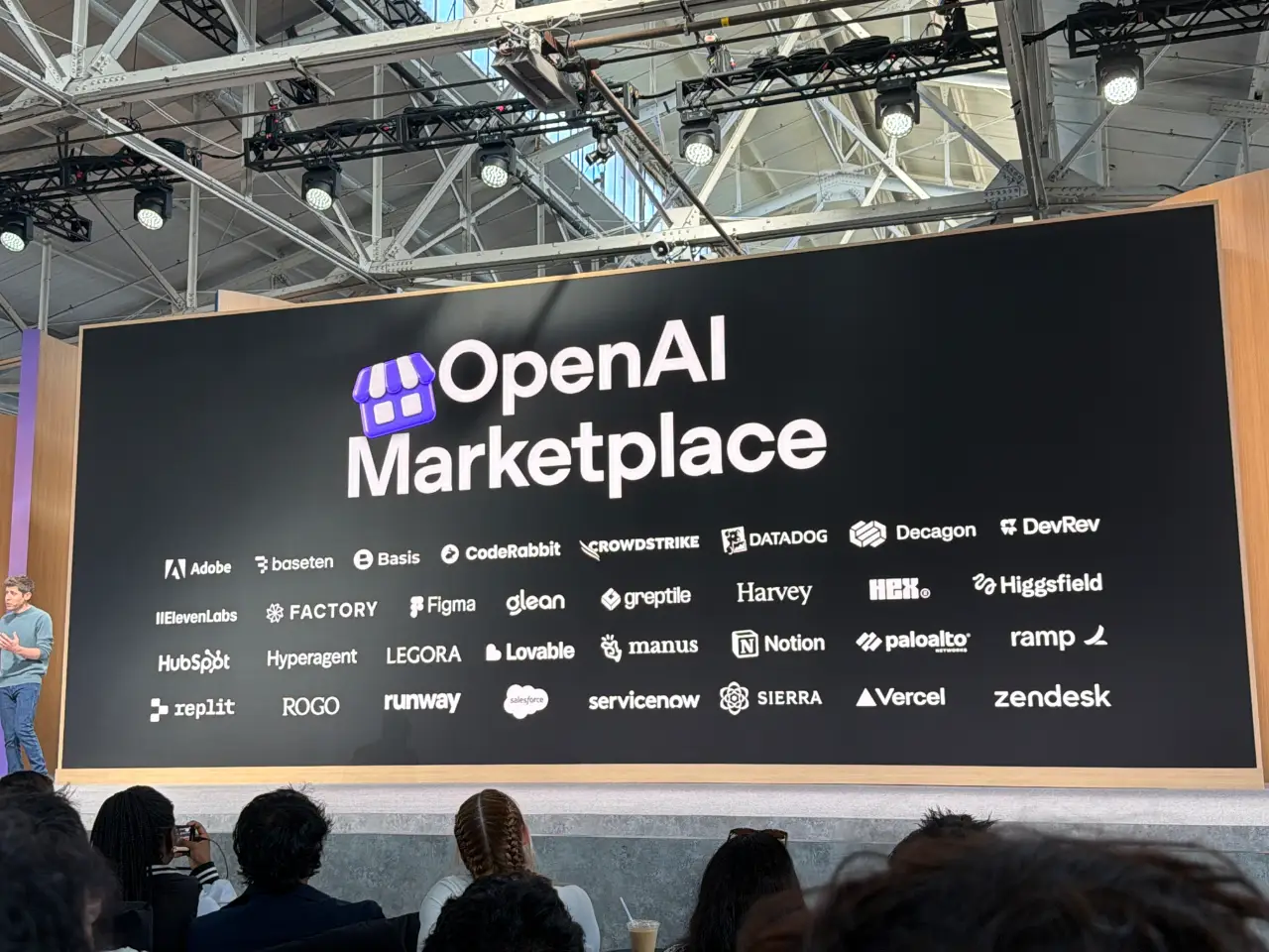 A large black stage display reads “OpenAI Marketplace,” with rows of logos including Adobe, Canva, Figma, Notion, Salesforce, Vercel and Zendesk. Audience members watch from the foreground beneath the venue’s exposed roof trusses and stage lights.