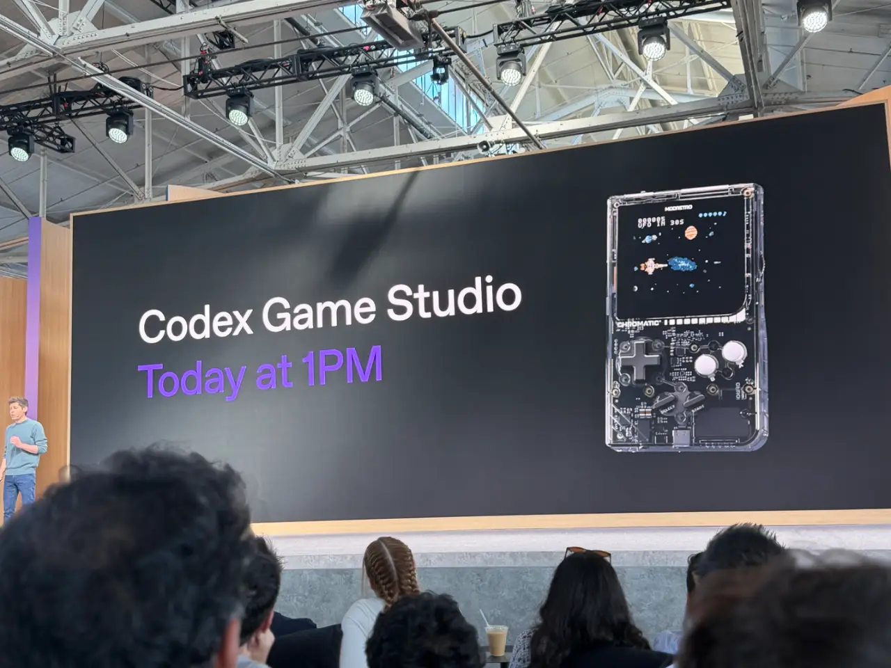 A presenter stands at the left of a stage before an audience, beside a large screen reading “Codex Game Studio” and “Today at 1PM,” with an image of a handheld game console.