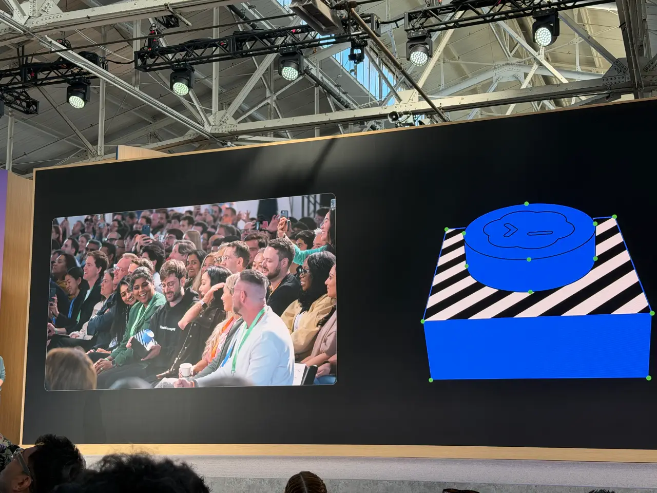 A large presentation screen at a conference shows an audience seated in rows on the left and a blue 3D graphic on the right: a rounded object with a cloud-like symbol sits on a black-and-white striped pedestal. Visible ceiling trusses and stage lights hang above, with attendees’ heads in the foreground.