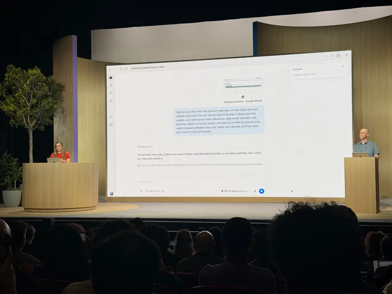 Two presenters stand at wooden podiums on either side of a large stage screen, addressing an audience seated in a dark auditorium. The screen shows a software interface titled “Build and publish launch radar,” with a prompt about turning a Google Sheet into a launch radar app.