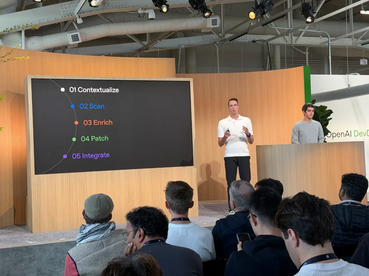 A presenter speaks to an audience at an OpenAI developer event, beside a large screen listing five steps: “01 Contextualize,” “02 Scan,” “03 Enrich,” “04 Patch” and “05 Integrate.” Another man stands behind a podium to the right.