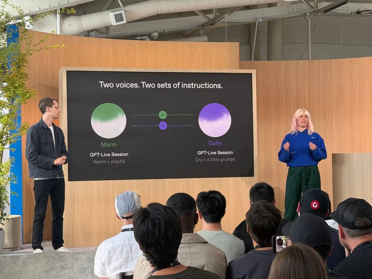 Two presenters address an audience on a wood-paneled stage beside a screen titled “Two voices. Two sets of instructions.” It compares GPT-Live Session voices Marin (“Warm + playful”) and Colin (“Dry + a little grumpy”) with green and purple graphics.