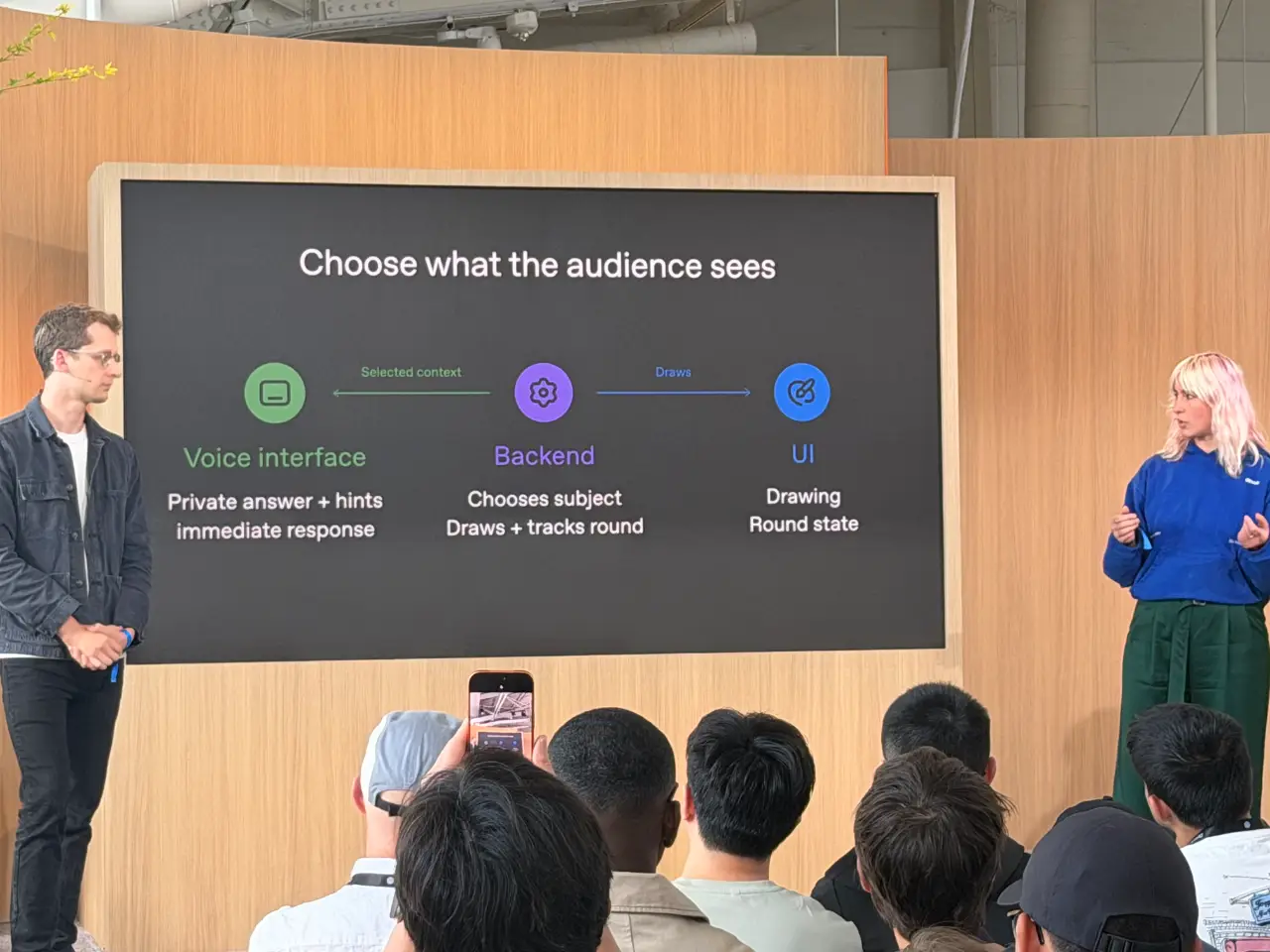 Two presenters stand beside a large screen during a talk, with an audience seated in the foreground. The slide, titled “Choose what the audience sees,” diagrams a voice interface, backend and UI, with notes about private answers, choosing a subject, drawing and tracking the round.