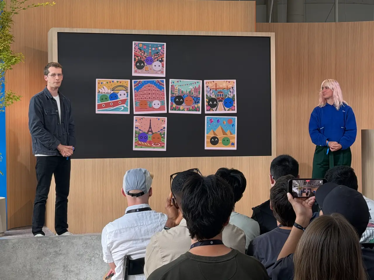 A man and a woman present to a seated audience in a wood-paneled room. Behind them, a large black display holds six colorful illustrations of cartoon faces against travel-themed backdrops; an attendee records the presentation on a phone.