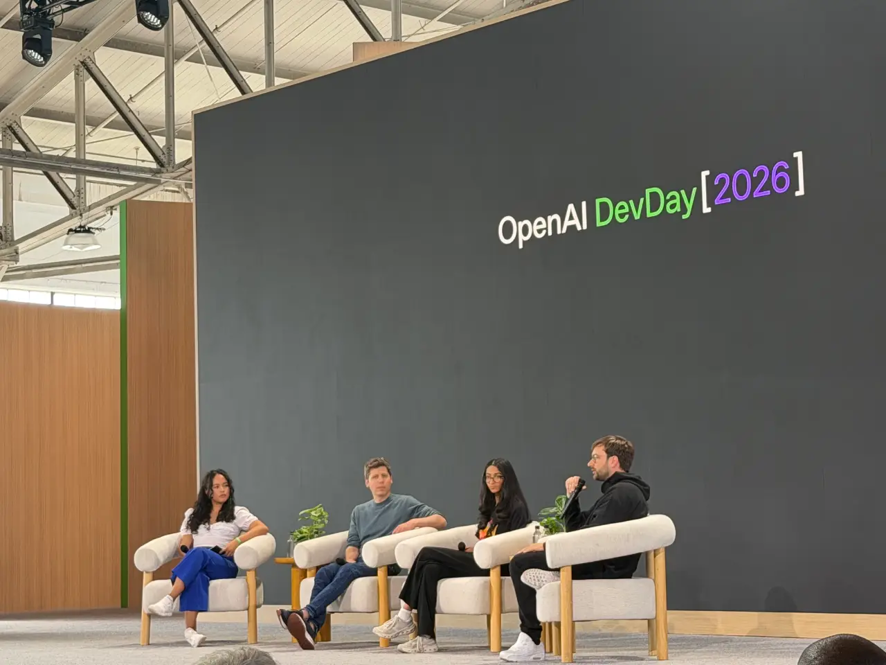 Four panelists sit in white armchairs on a stage beneath a large screen reading “OpenAI DevDay [2026].” The man on the right speaks into a microphone while the others listen.
